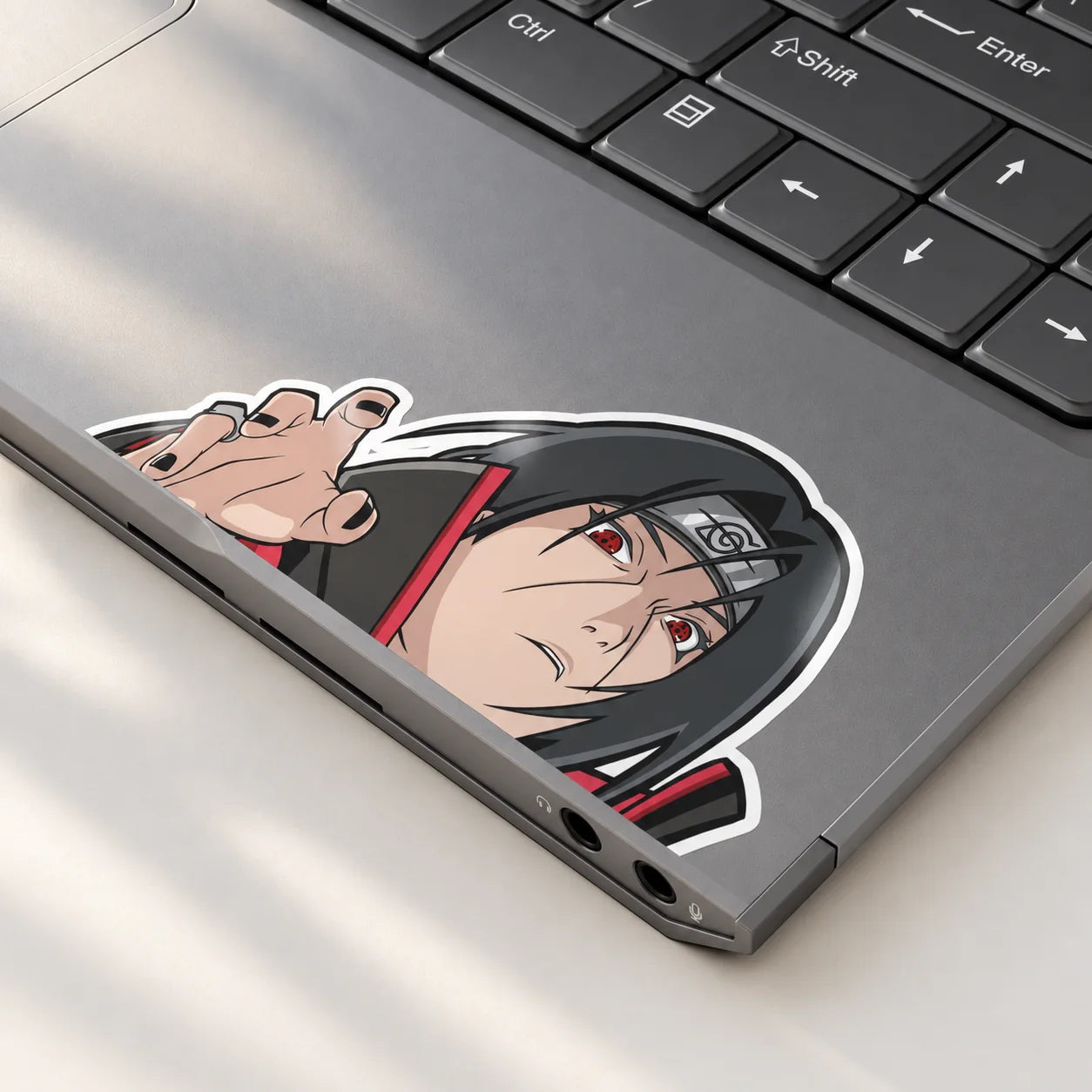 Itachi peeker vinyl sticker applied flush with the edge of a grey laptop, showing its glossy finish